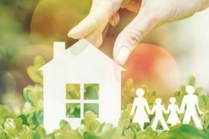 іграшковий паперовий будиночок та іграшкова паперова сім'я біля нього, рука ставить їх на траву