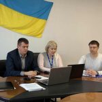 люди сидять за столом, прапор України на стіні за ними