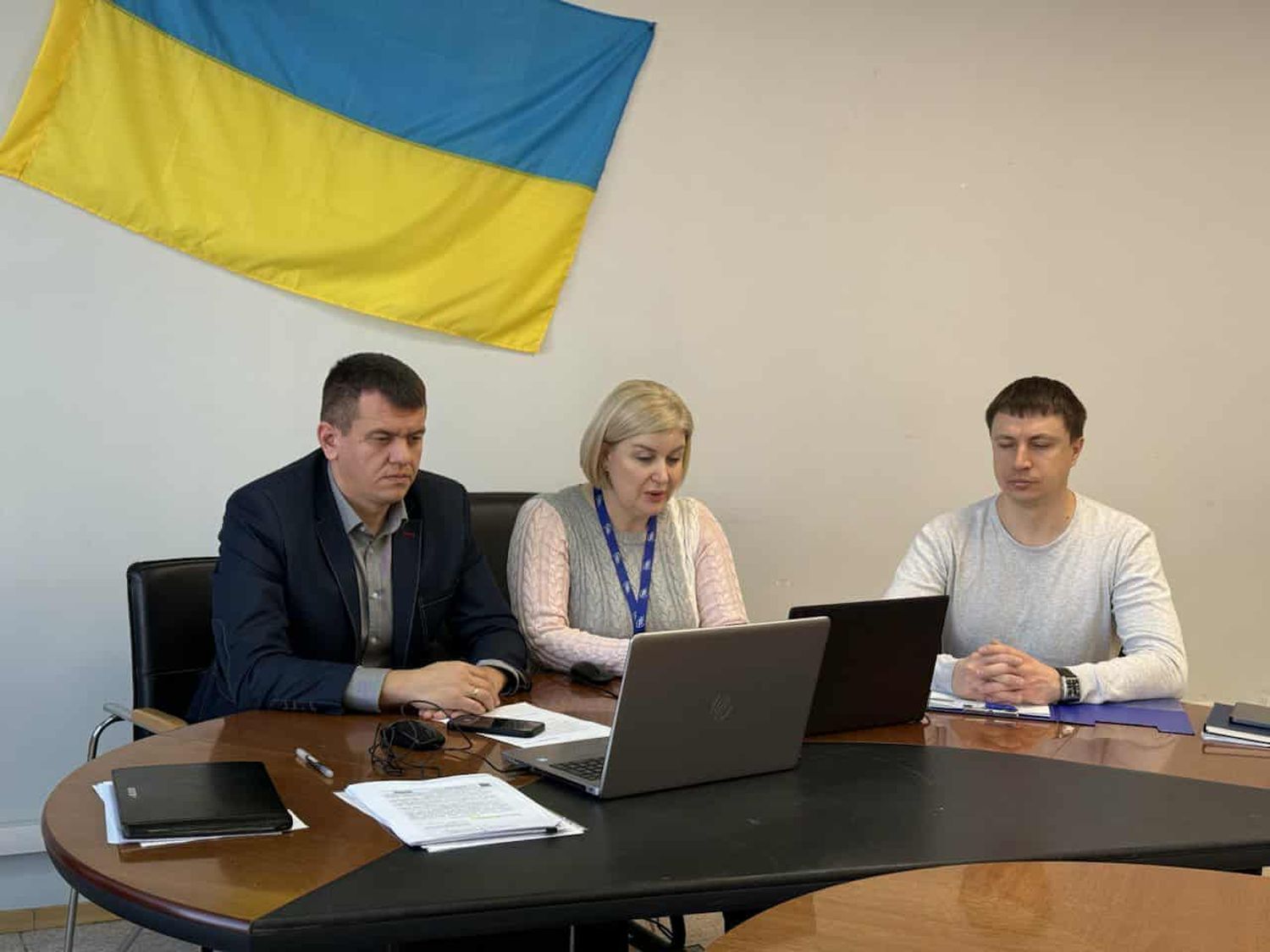 люди сидять за столом, прапор України на стіні за ними